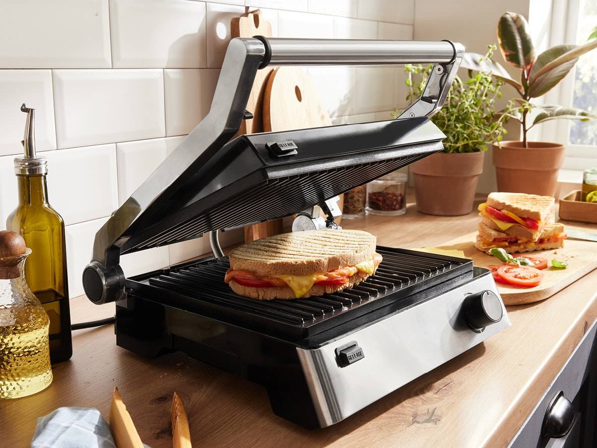 Offener Sandwichmaker mit einem Käse-Tomaten-Toast, weitere Toasts auf einem Schneidebrett.