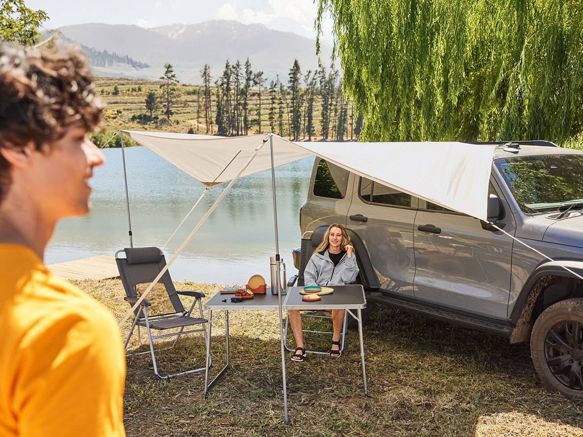 Coppia in campeggio con tenda da sole per auto, tavolo pieghevole e sedie da campeggio vicino a un lago.