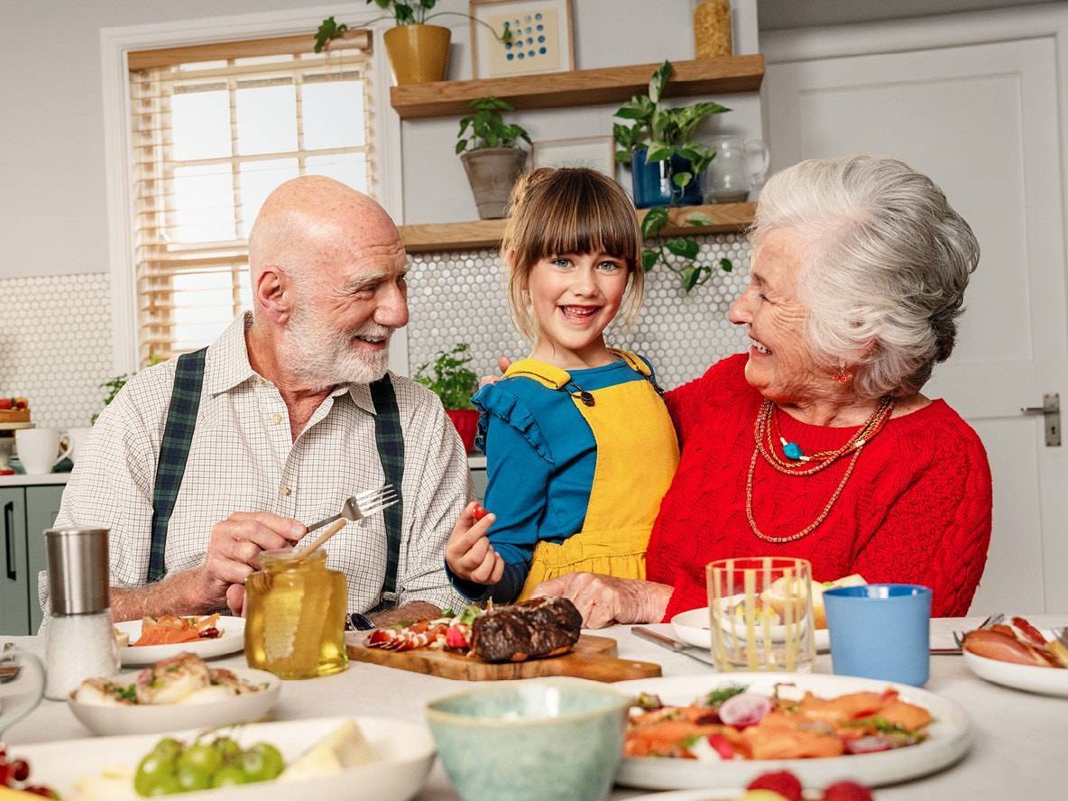 Nonni e nipote sorridenti a tavola, con salmone affumicato, carne e frutta.