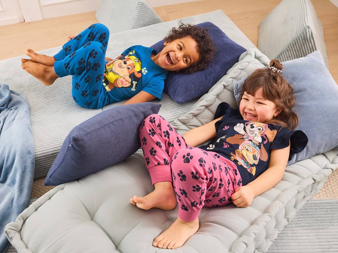 Deux enfants rient allongés sur des coussins de sol, vêtus de pyjamas à motifs de pattes.