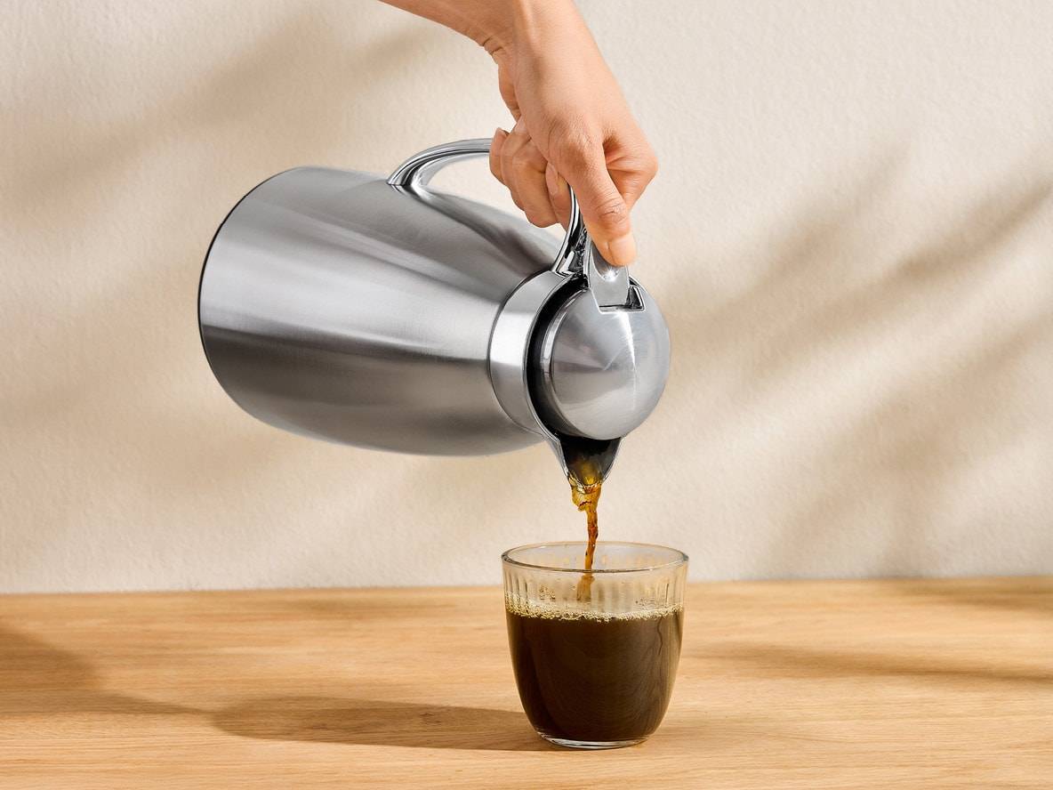 Une main verse du café noir d'une carafe isotherme en acier inoxydable dans un verre.