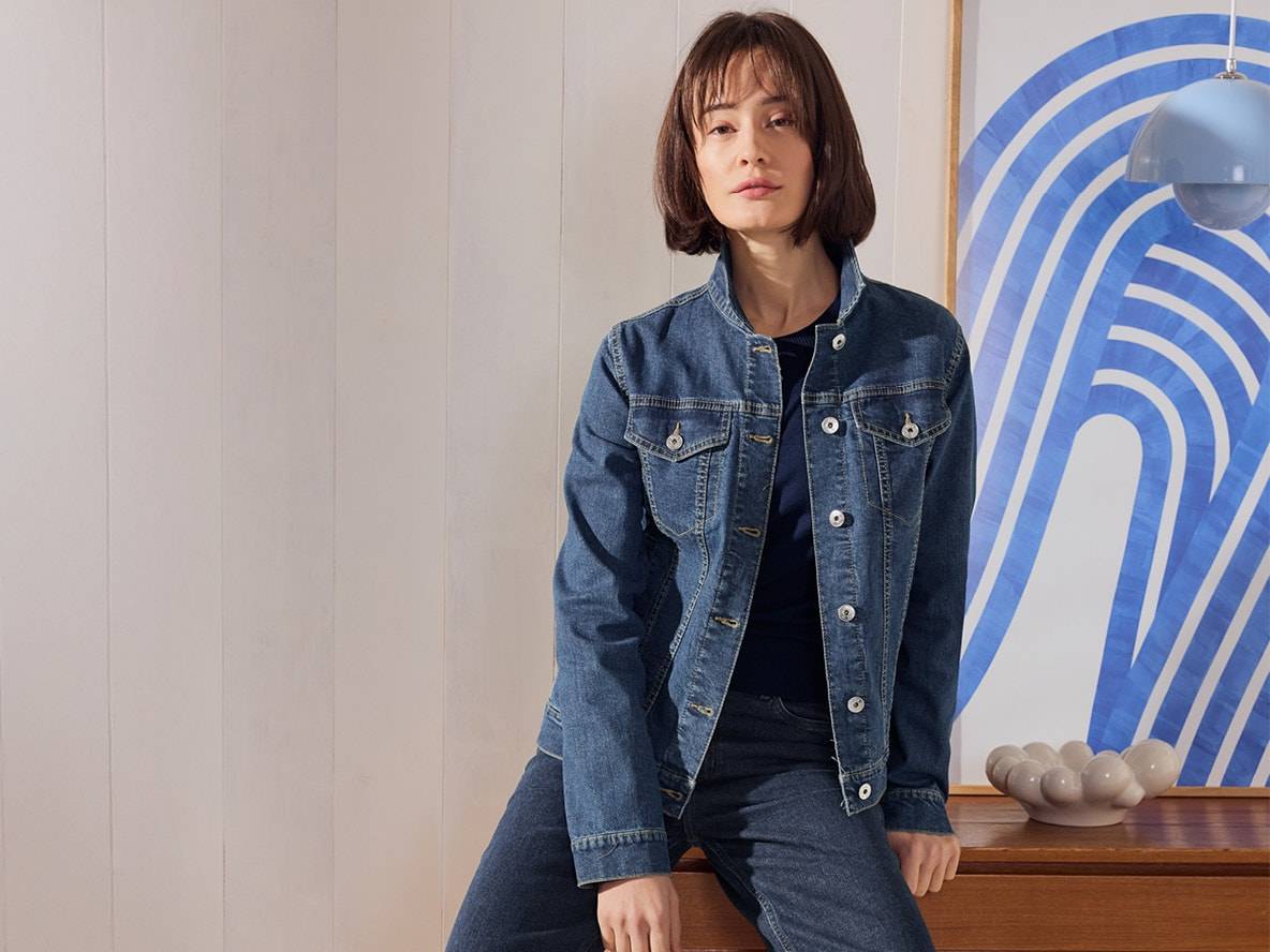Donna con giacca e jeans di denim, seduta su un mobile di legno.