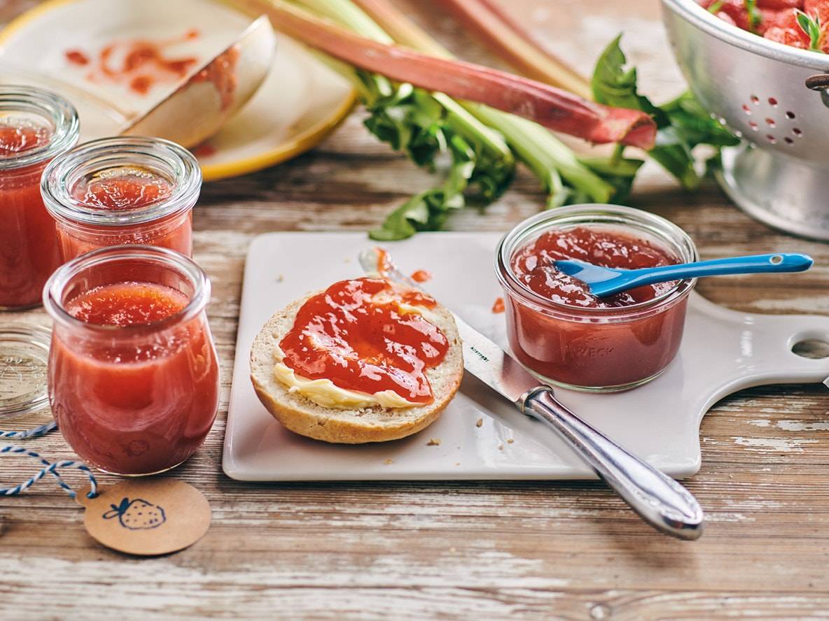 Confiture de fraises et rhubarbe en pots, avec un petit pain tartiné et de la rhubarbe fraîche.