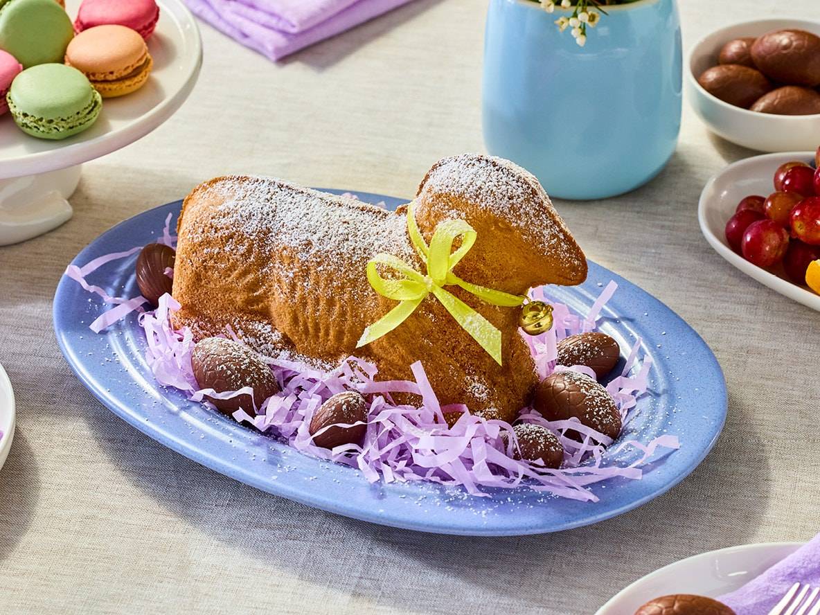 Torta a forma di agnello pasquale con zucchero a velo, cioccolatini a forma di uovo e macarons colorati.
