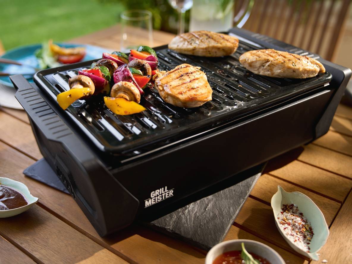 Elektrogrill mit Gemüsespießen und gegrillten Hähnchenbrustfilets auf einem Holztisch im Freien.