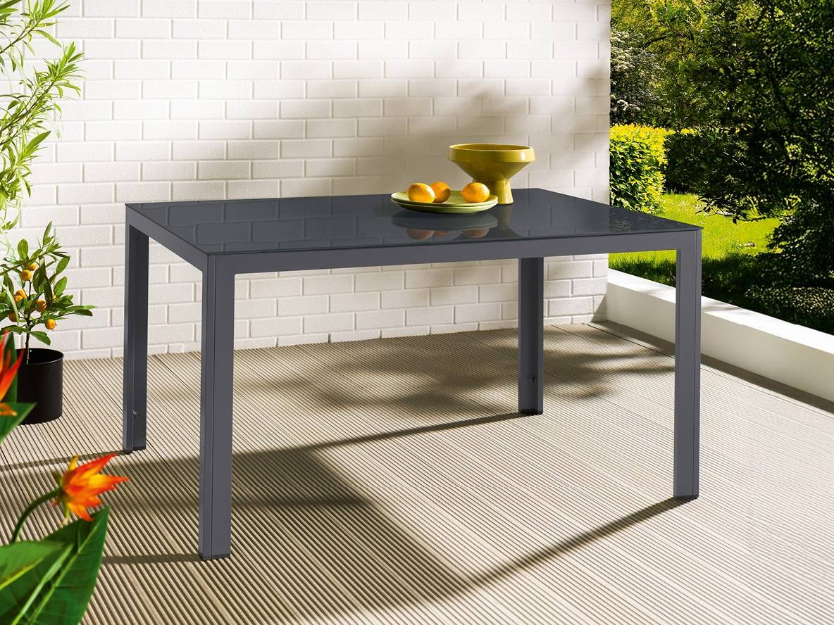 Grauer Gartentisch mit Glasplatte auf einer Terrasse mit Pflanzen und Garten im Hintergrund.