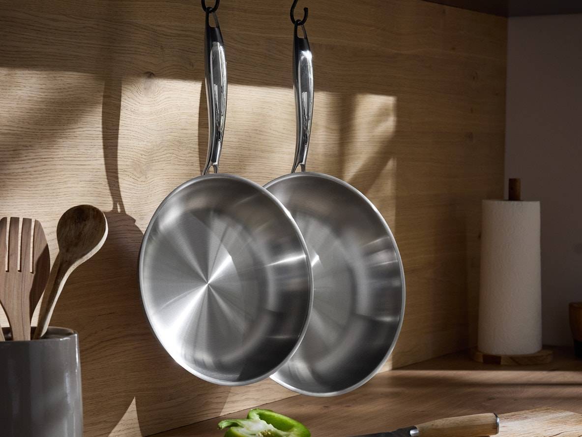 Deux poêles en acier inoxydable suspendues à un mur en bois dans une cuisine, avec des ustensiles en bois et un rouleau de papier.