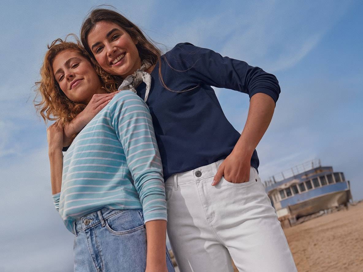 Zwei Frauen in Langarmshirts und Jeans, mit einem gestrandeten Schiff im Hintergrund.
