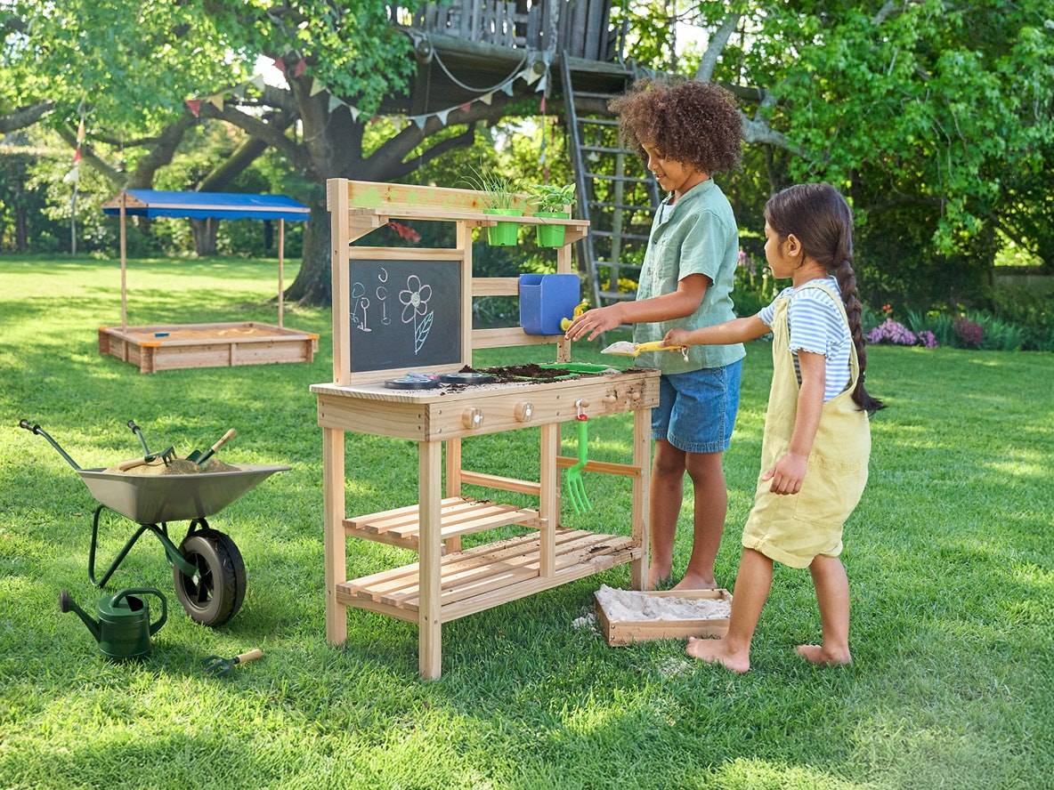 Zwei Kinder spielen mit einem Holzgärtnertisch und einer Schubkarre im Garten.