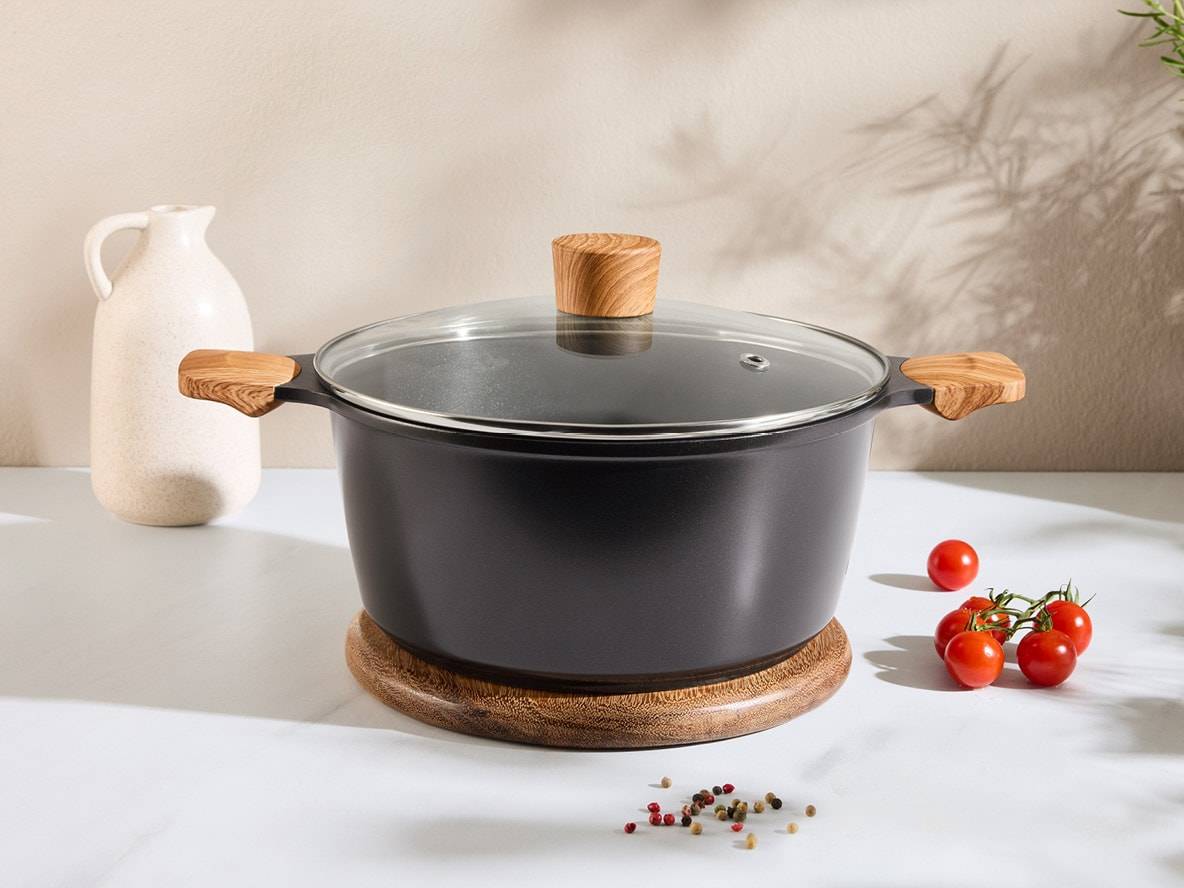 Schwarzer Topf mit Glasdeckel und Holzgriffen, auf einem Holzuntersetzer, mit Tomaten und Gewürzen.