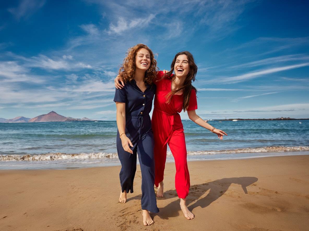 Zwei Frauen in blauen und roten Overalls gehen lachend am Strand entlang.