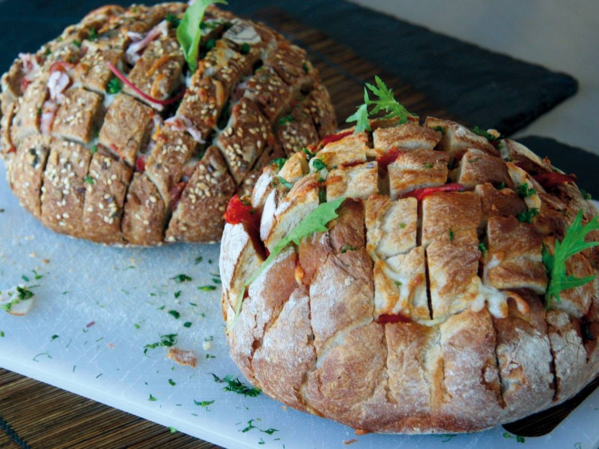 Zwei gefüllte Ziehharmonikabrote mit Käse und Gemüse auf einem Schneidebrett.