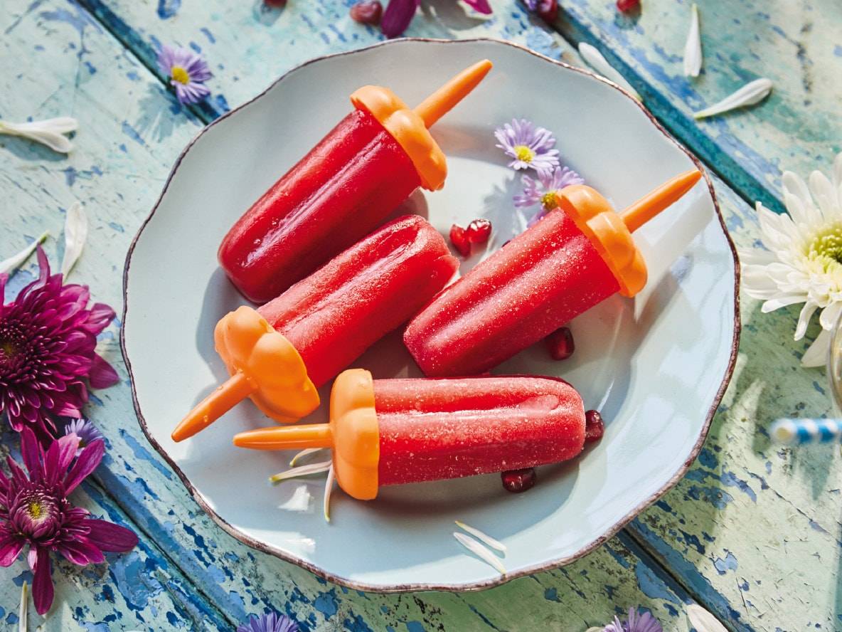 Vier rote Eis am Stiel auf einem blauen Teller, dekoriert mit Blumen und Granatapfelkernen.