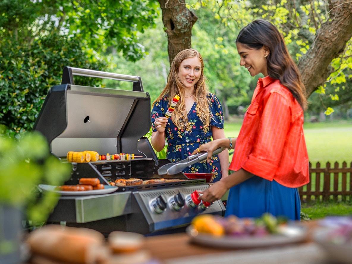 Zwei Frauen grillen im Garten, mit Spießen und Würstchen auf dem Grill.