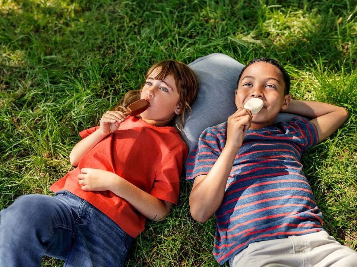 Zwei Kinder liegen im Gras und essen Eis am Stiel.
