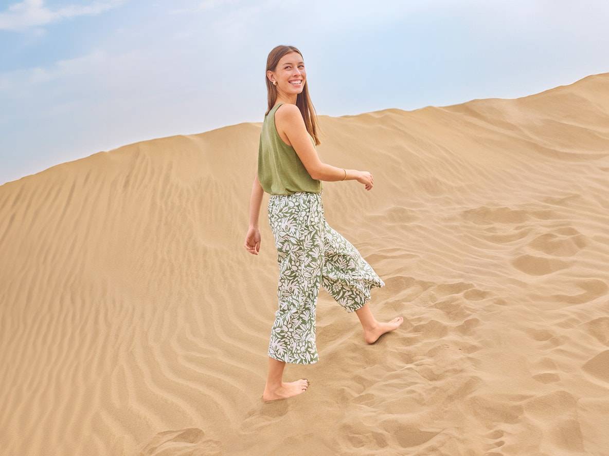 Frau in grünem Top und gemusterter Hose geht barfuß durch Sanddünen.