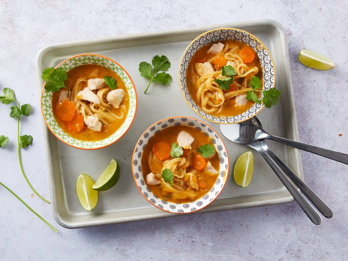 Drei Schalen Hühnernudelsuppe mit Karotten und Koriander auf einem Tablett, garniert mit Limettenspalten.