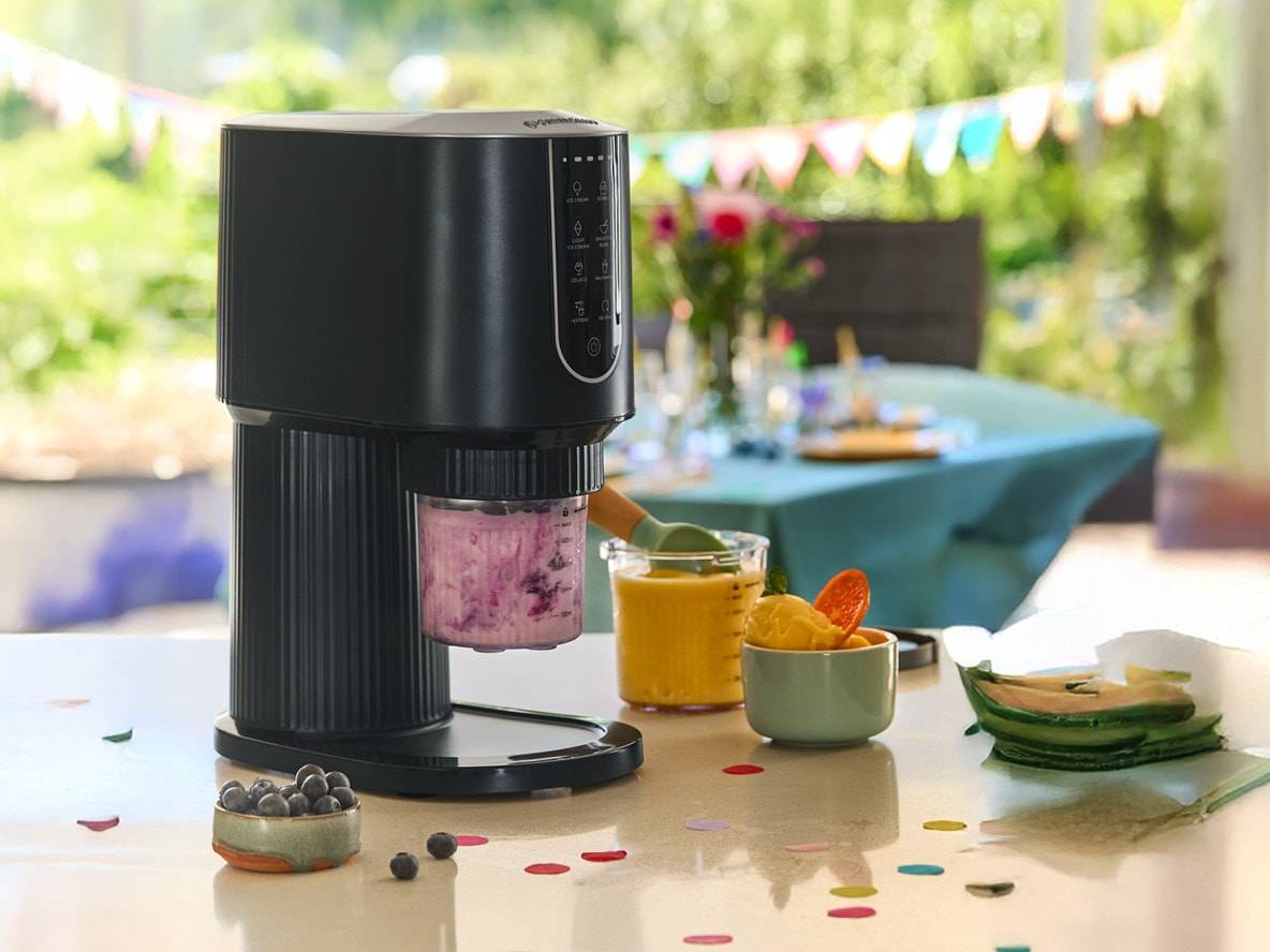 Eismaschine mit lila Eiscreme, daneben gelbes Sorbet und Blaubeeren auf einem Tisch mit Konfetti.