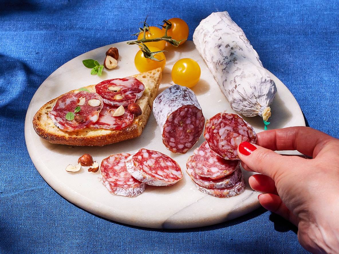 Fette di salame e salame intero su un tagliere di marmo con pane, nocciole e pomodori.