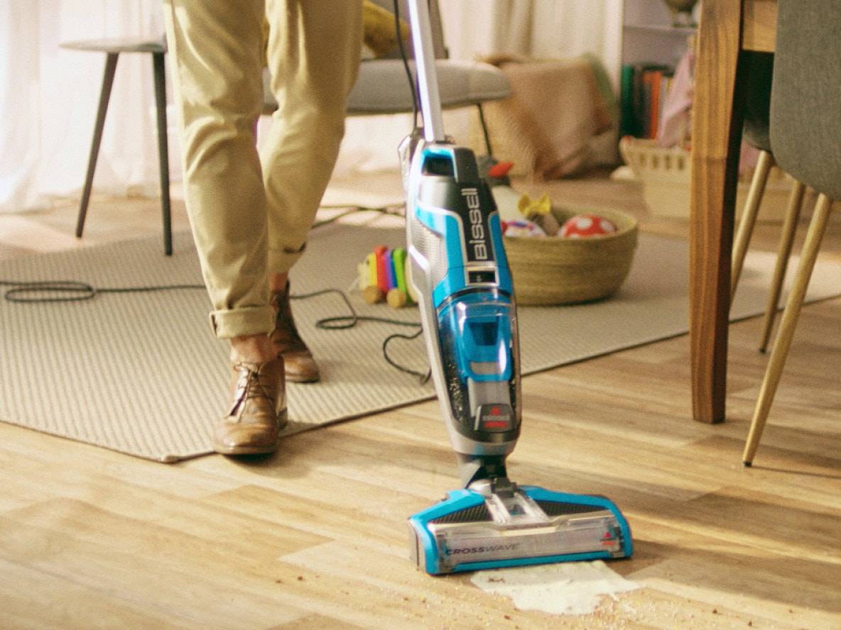 Aspirateur balai nettoyant une tache sur un sol en bois, personne debout.