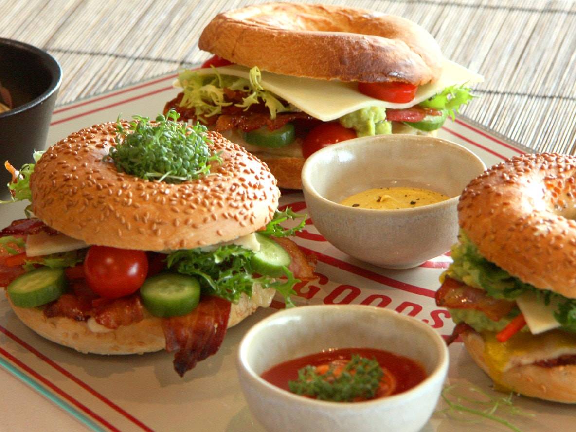 Drei Bagel-Sandwiches mit verschiedenen Füllungen und Dips auf einem Tablett.