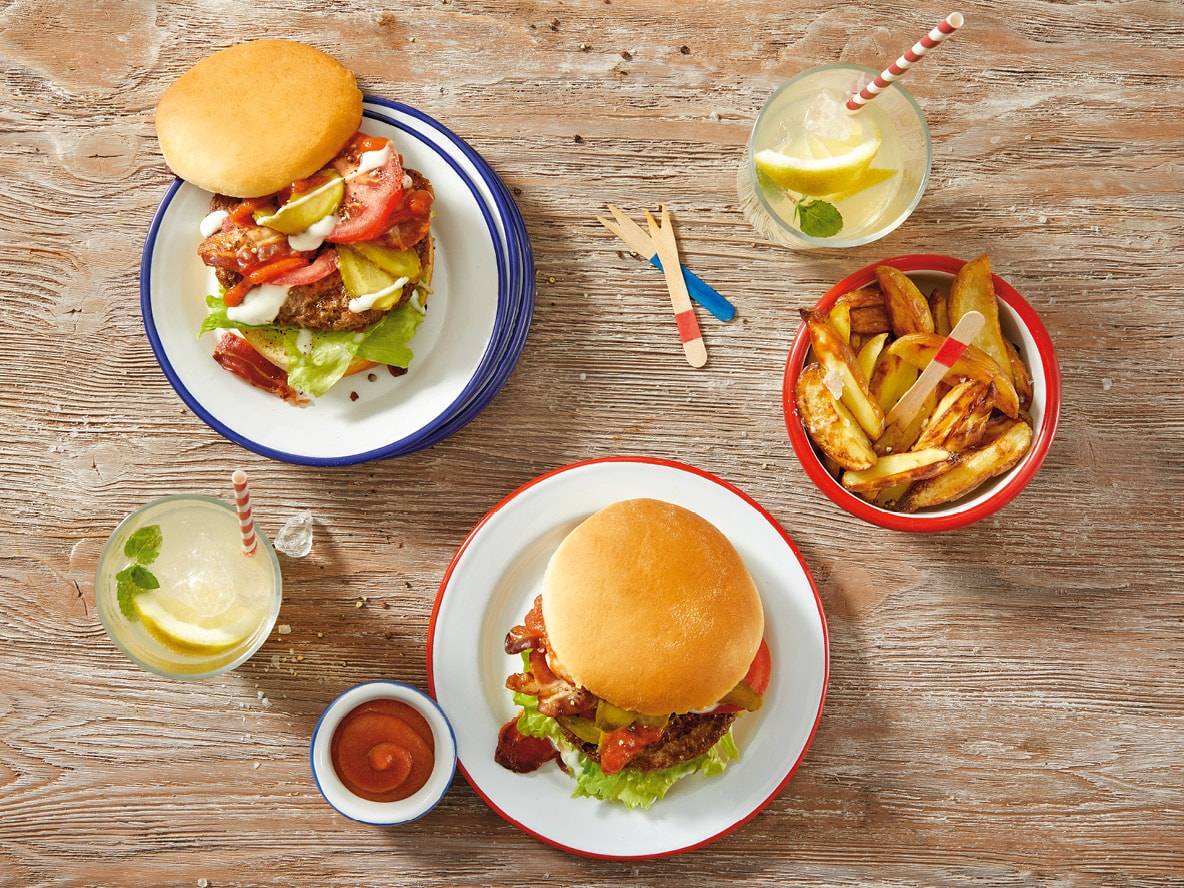 Zwei Burger mit Pommes Frites und Limonade auf einem Holztisch.