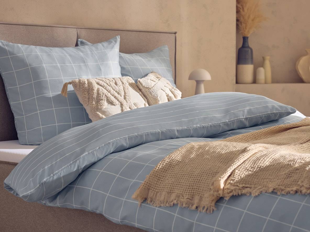 Bett mit hellblauer Bettwäsche mit Karomuster, Zierkissen und beiger Decke.