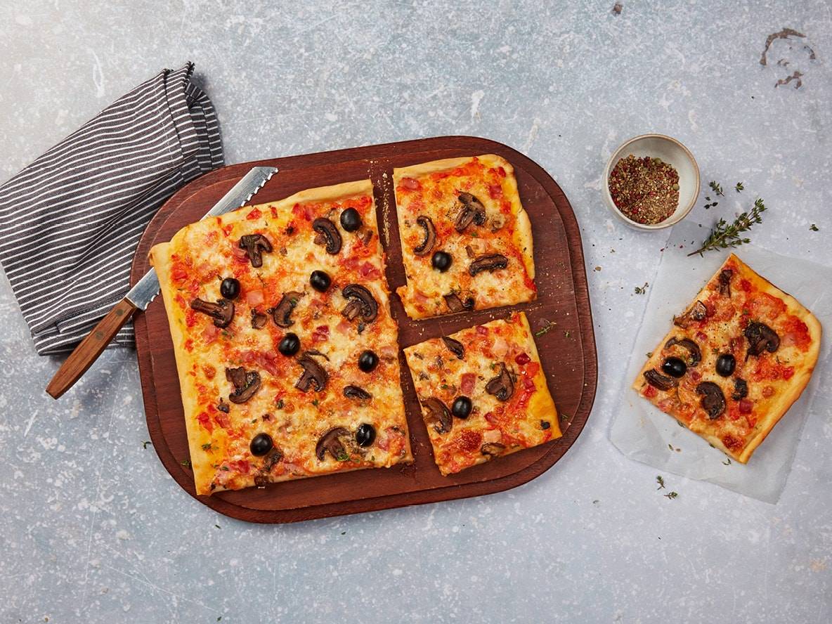 Rechteckige Pizza mit Pilzen und Oliven auf einem Holzbrett, mit einem separaten Stück.