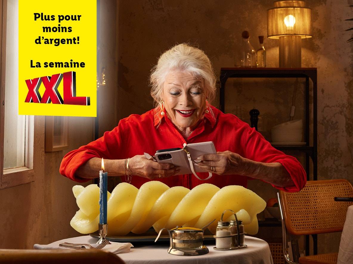 Une femme âgée utilise son téléphone portable à côté d'une grande portion de pâtes XXL, faisant la promotion d'une offre spéciale.