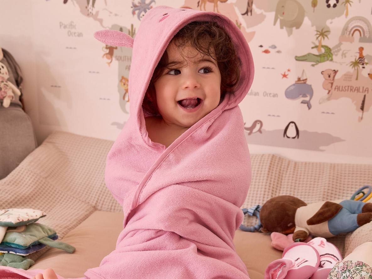 Un jeune enfant enveloppé dans un peignoir rose à capuche est assis sur un lit.