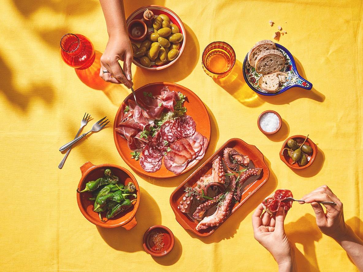 Table de tapas avec charcuterie, poulpe, poivrons, olives et pain sur nappe jaune.