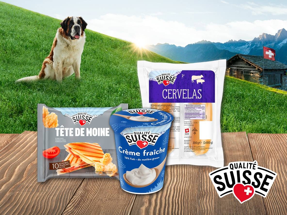 Qualité Suisse Produkte (Cervelas, Tête de Moine, Crème fraîche) auf einem Holztisch mit einem Bernhardiner und Schweizer Berglandschaft.