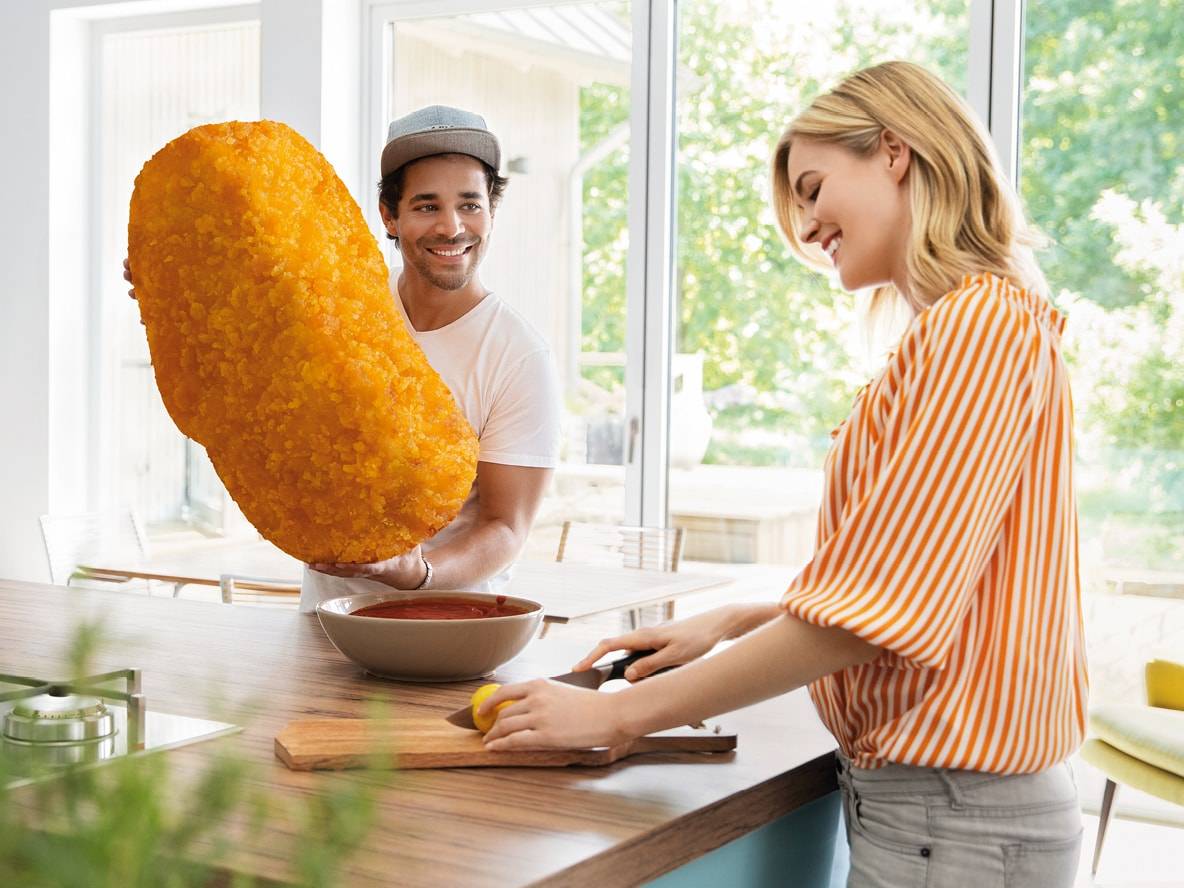 Ein lächelnder Mann hält ein riesiges Chicken Nugget, während eine Frau eine Zitrone schneidet.