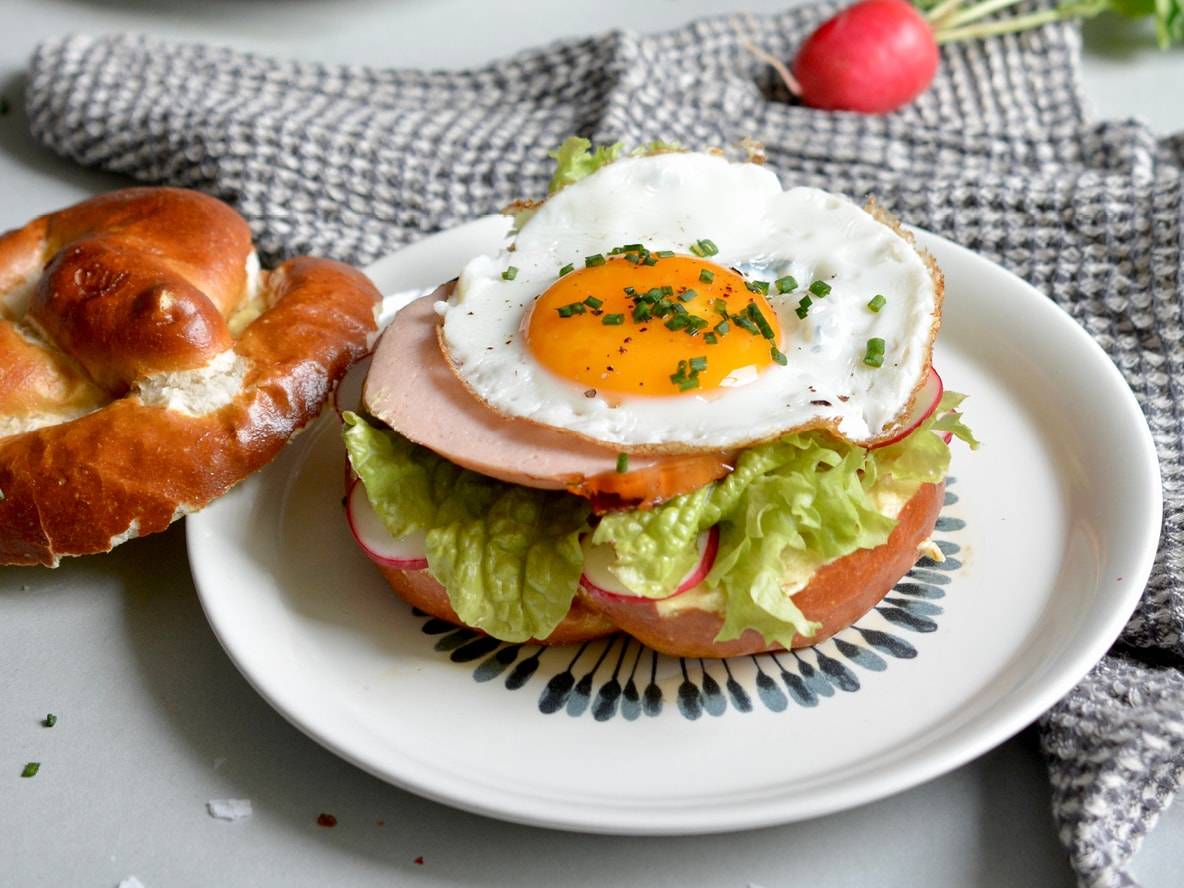 Ein Brezel-Sandwich mit Spiegelei, Salat, Radieschen und Fleisch, mit einer Radieschen im Hintergrund.