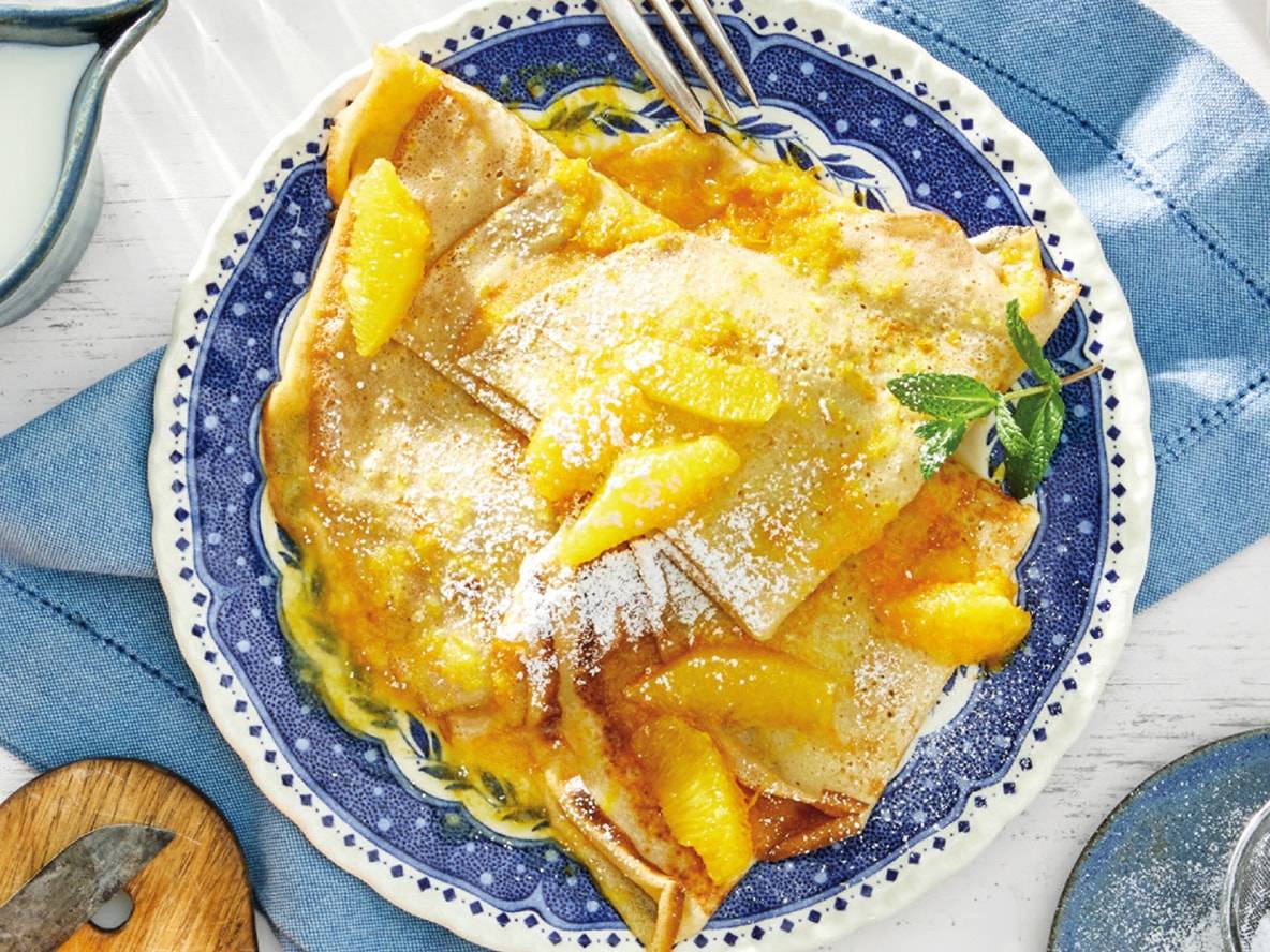 Orangen-Crêpes mit Puderzucker und Minze auf einem blau-weißen Teller.