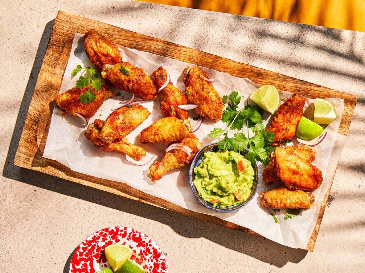 Gebratene Hähnchenflügel mit Guacamole, Limetten und roten Zwiebeln auf einem Holzbrett.