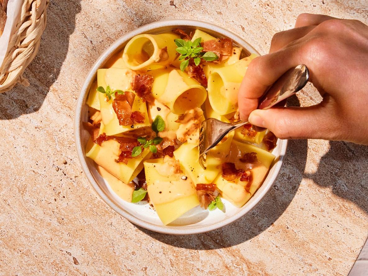 Eine Hand isst mit einer Gabel eine Schüssel Pasta mit knusprigem Speck und Basilikum auf einer Steinoberfläche.