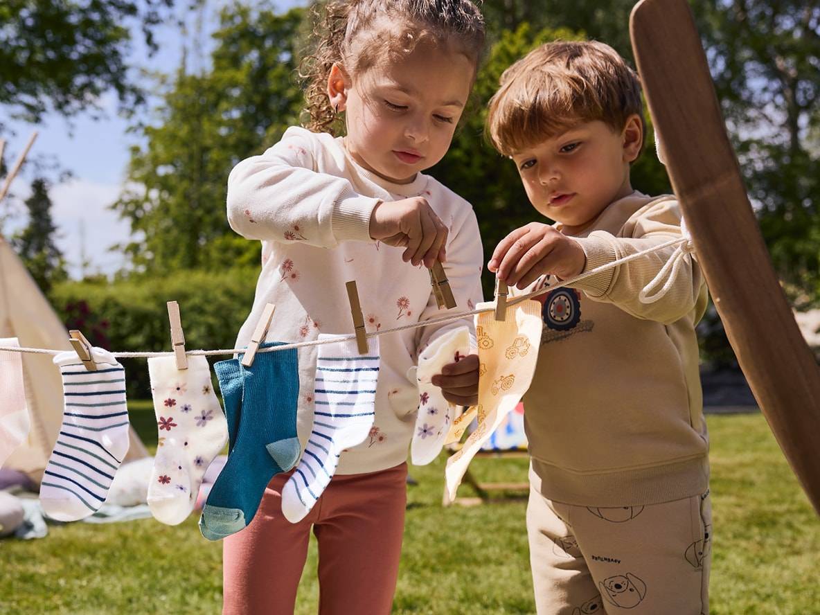 Zwei Kinder hängen Socken und Stoffe auf eine Wäscheleine im Freien.