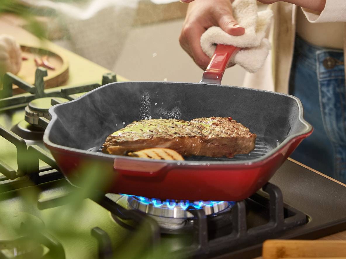 Eine Person brät ein Steak in einer roten Pfanne auf einem Gasherd.