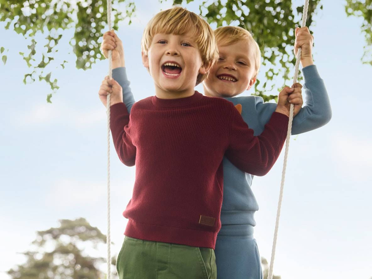 Deux jeunes garçons souriants sur une balançoire, portant des pulls et des pantalons décontractés.