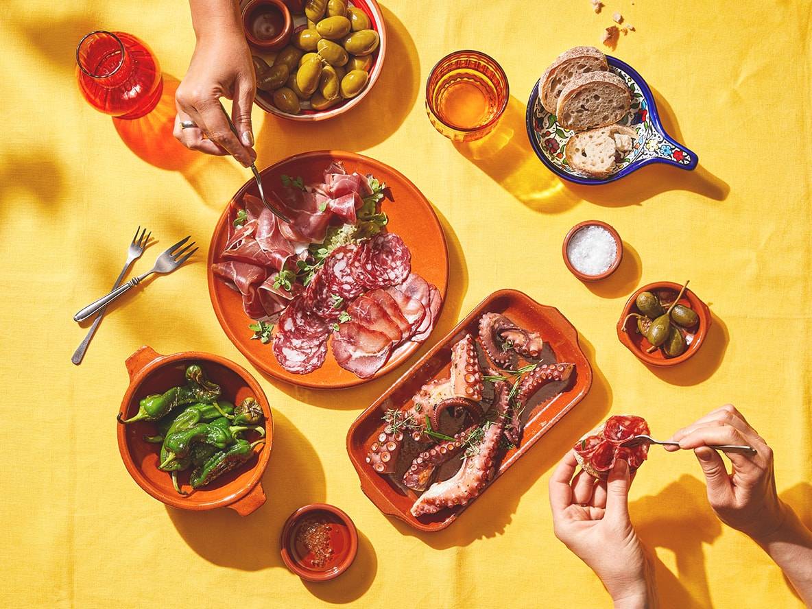 Une table jaune avec des tapas espagnoles, incluant charcuterie, poulpe, poivrons et olives.