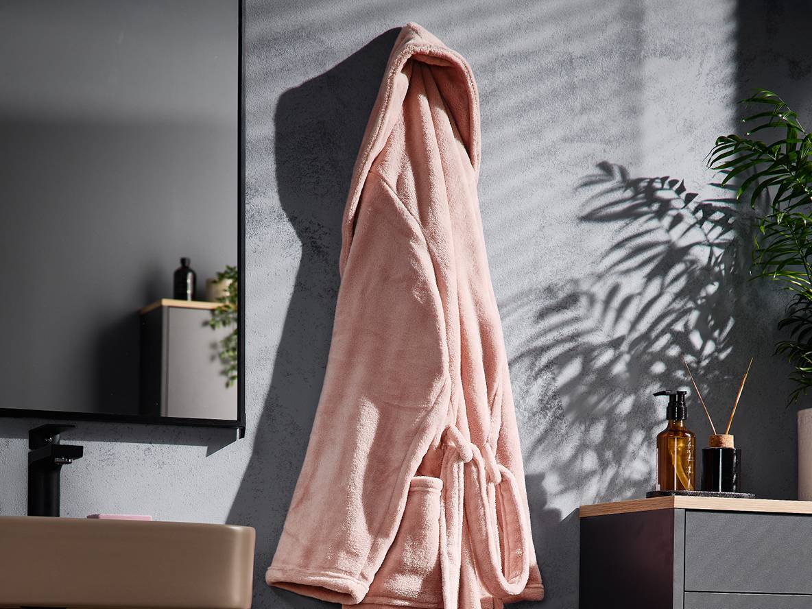 Rosa Bademantel hängt an einer strukturierten Badezimmerwand mit Spiegel und Waschbecken.