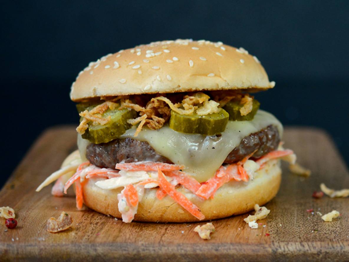 Ein Burger mit Krautsalat, Gurken, Käse und Röstzwiebeln auf einem Holzbrett.