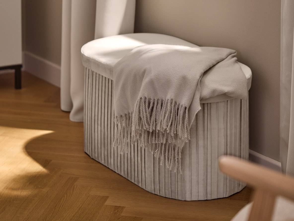 Panca contenitore in velluto beige con coperta, su pavimento in legno.