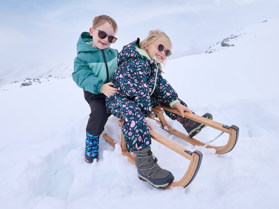 Zwei lächelnde Kinder in Schneeanzügen und Sonnenbrillen, eines auf einem Holzschlitten.