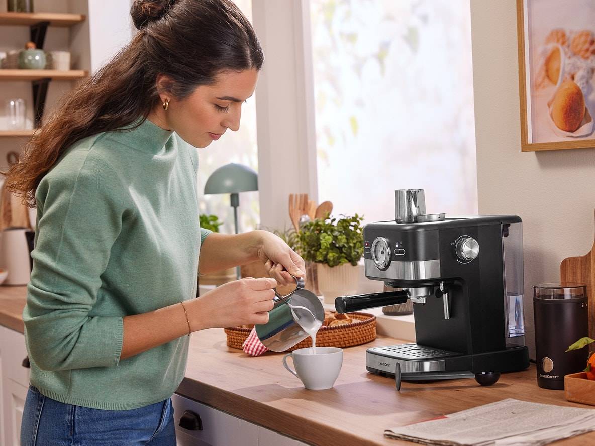 Frau gießt Milch in eine Tasse, mit einer SilverCrest Espressomaschine auf der Theke.