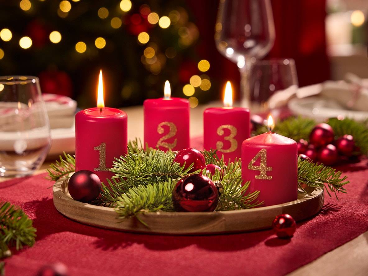 Roter Adventskranz mit vier nummerierten Kerzen, geschmückt mit Tannenzweigen und Kugeln.