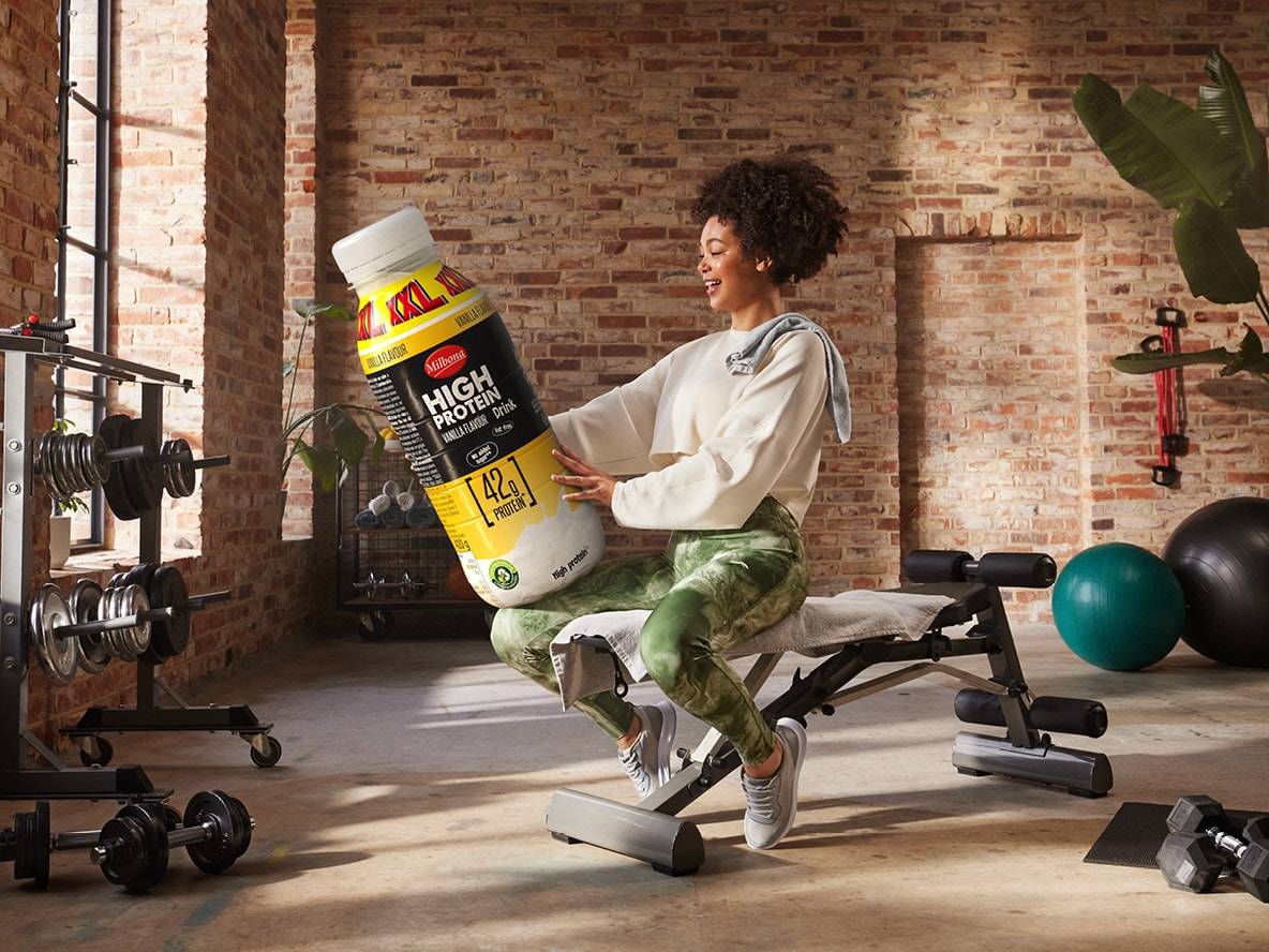 Eine lächelnde Frau hält eine große Milbona Protein-Drinkflasche in einem Fitnessstudio.