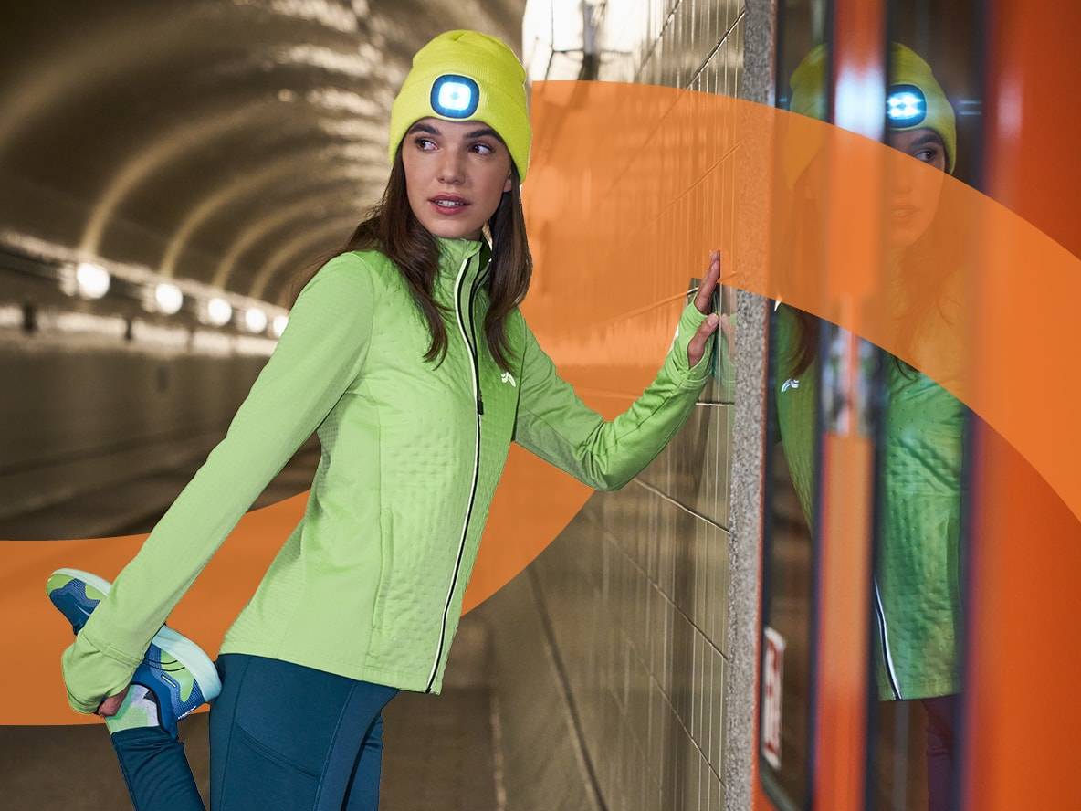 Frau in Crivit Sportkleidung, Mütze mit Stirnlampe, Jacke und Leggings, dehnt sich in einem Tunnel.