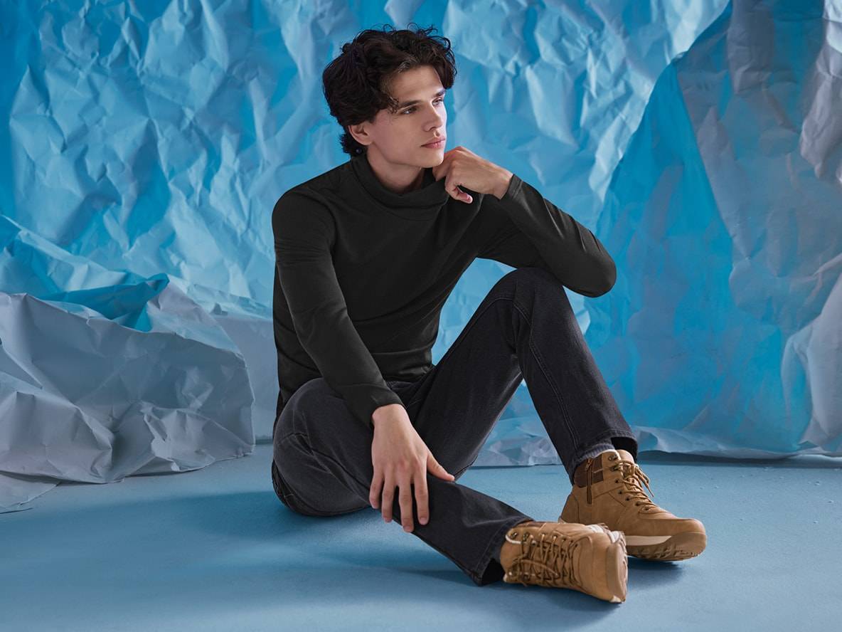 Mann in schwarzem Rollkragenpullover, dunklen Jeans und braunen Stiefeln, sitzend vor blauem Hintergrund.